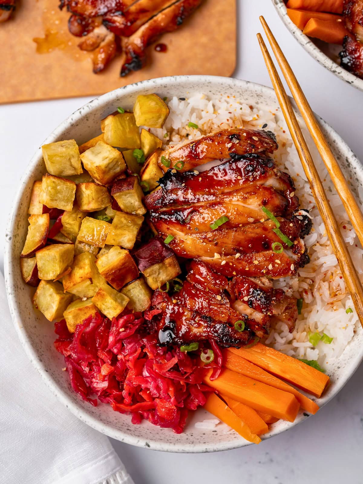 sliced air fried chicken thigh in a bowl with steamed rice, roasted japanese sweet potatoes, pickled cabbage and carrots, and toasted sesame seeds