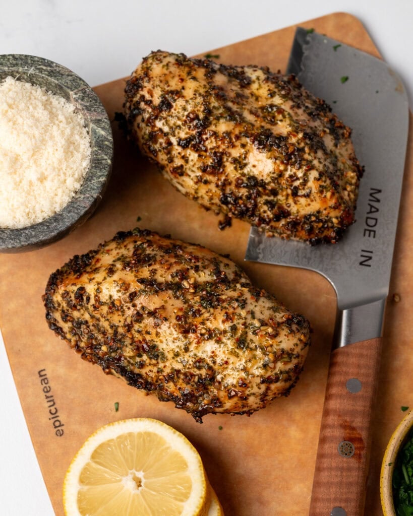 two chicken breasts on a cutting board next to chopped parsley, lemon slices, and grated cheese