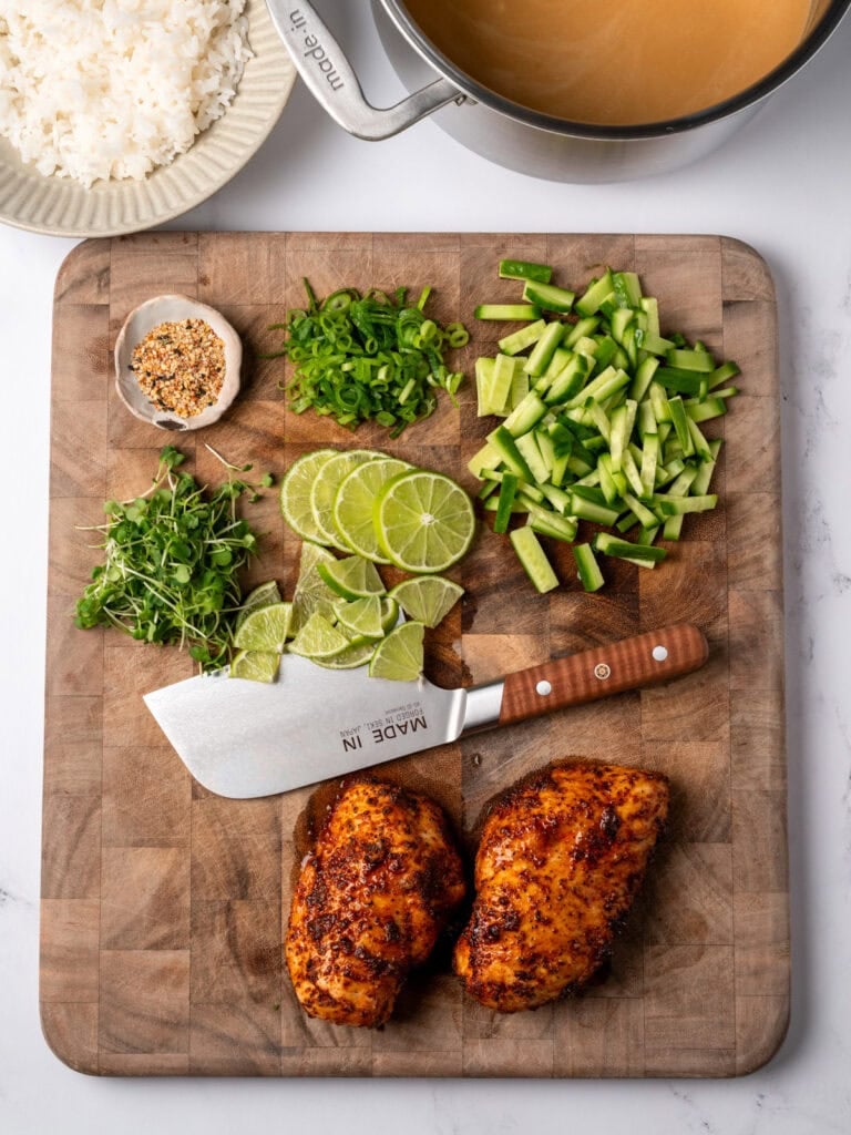 chicken breasts resting on a cutting board next to sliced cucumbers, scallions, baby bok choy, and lime wedges