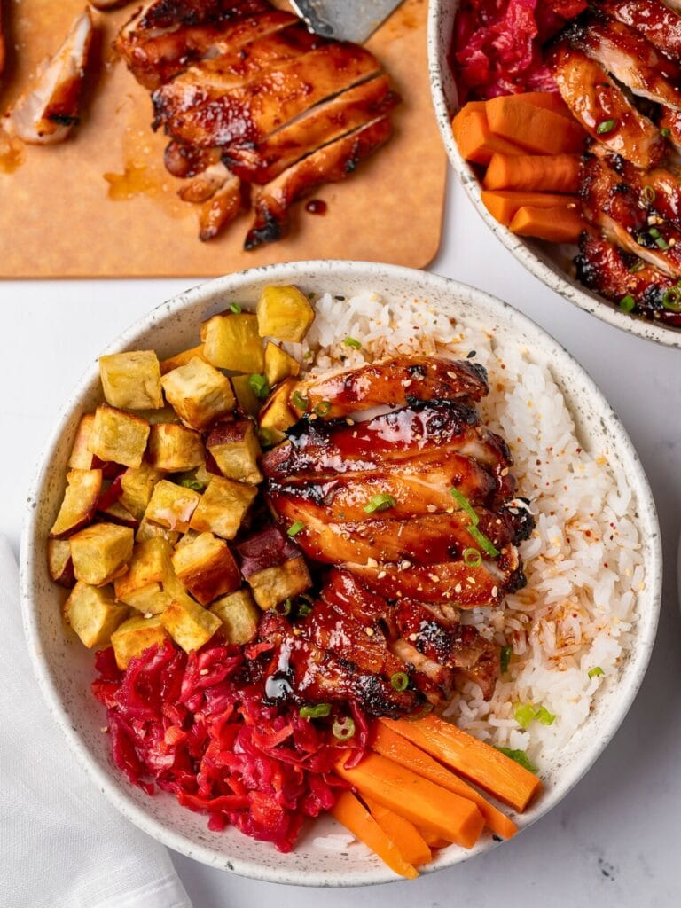 sliced Japanese bbq chicken thigh in a bowl with steamed rice, roasted japanese sweet potatoes, pickled cabbage and carrots, green onion, and toasted sesame seeds