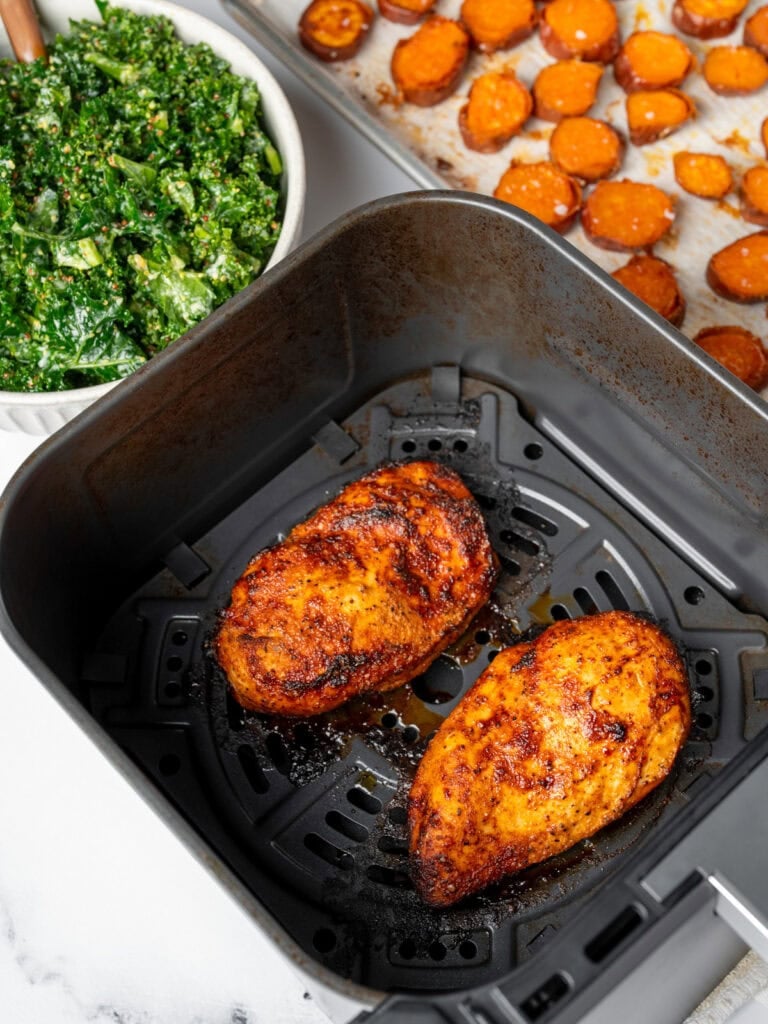 two air fried chicken breasts in an air fryer basket next to kale salad and roasted sweet potatoes on a sheet pan