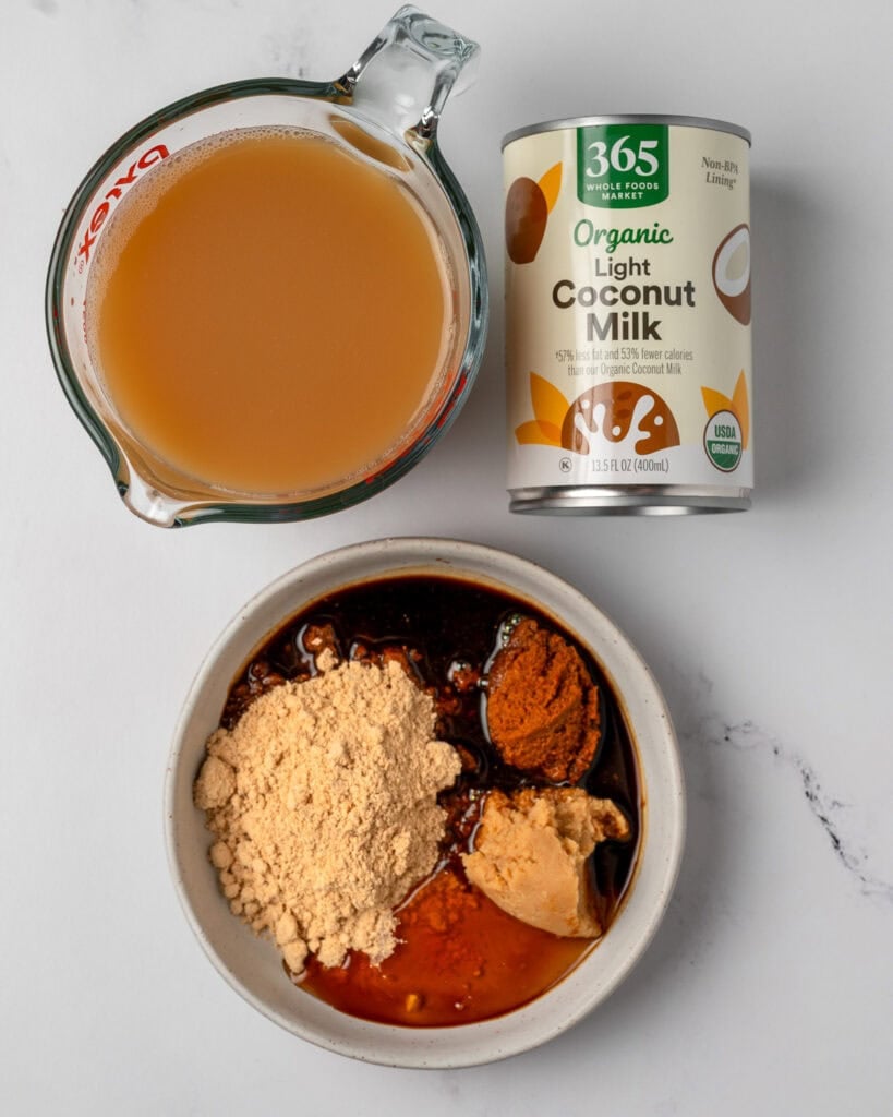 a small bowl with powdered peanut butter, yellow curry paste, white miso paste, honey, and coconut aminos next to a can of light coconut milk and a measuring cup with chicken broth
