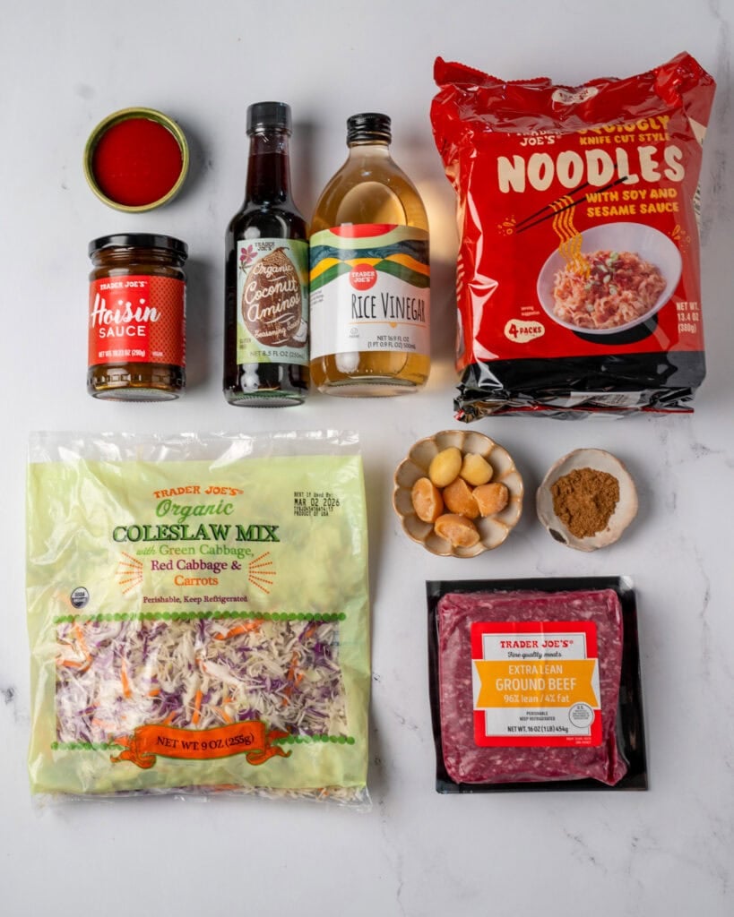 Trader Joe's knife cut squiggly noodles, rice vinegar, coconut aminos, hoisin sauce, sriracha, coleslaw mix, and extra lean ground beef next to frozen ginger and garlic cubes and Chinese five spice