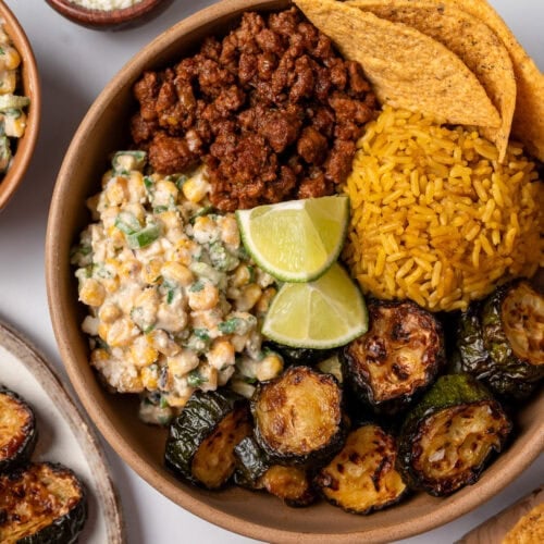 a brown bowl with taco beef, street corn salad, roasted zucchini, Spanish rice, tortilla chips, and lime wedges