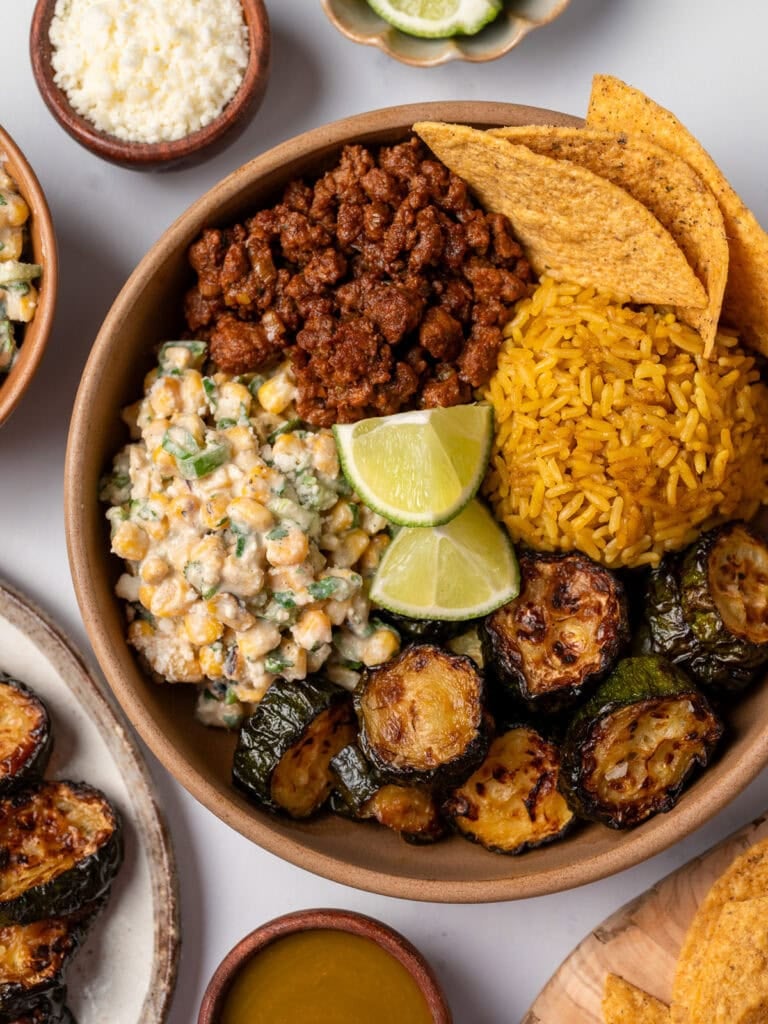 a brown bowl with taco beef, street corn salad, roasted zucchini, Spanish rice, tortilla chips, and lime wedges