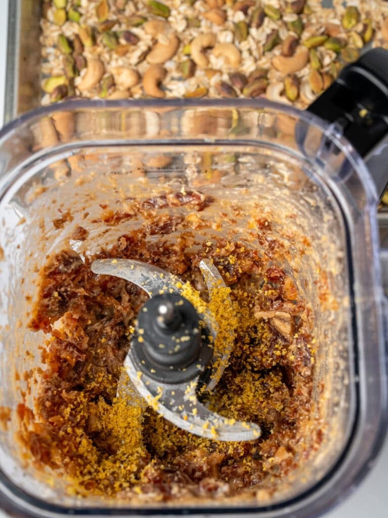 blended dates, cashew butter, vanilla, honey, and lemon zest in a blender next to a sheet pan with toasted oats, cashews, and pistachios