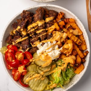 ground beef and air fried butternut squash zig zags in a bowl with lettuce, pickles, diced tomatoes, crumbled cheese, and burger sauce
