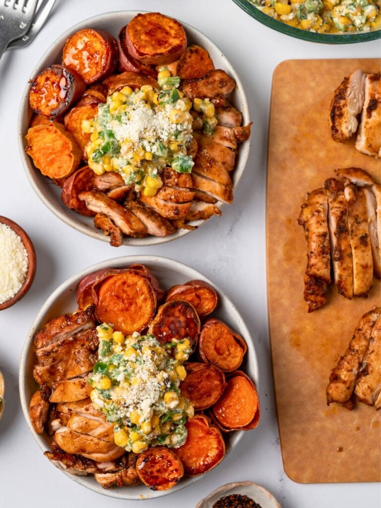 two esquites bowls with roasted sweet potatoes and chicken next to a cutting board with sliced chicken and garnishes like cotija cheese