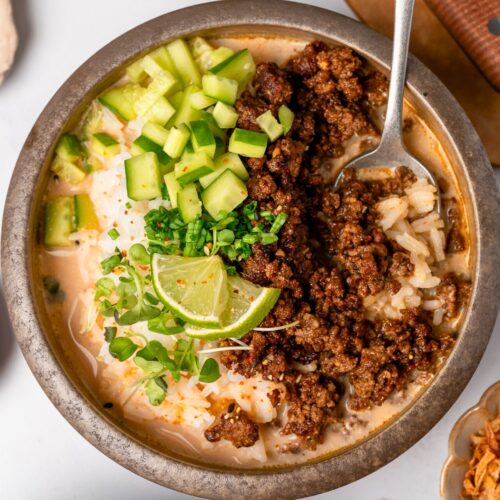 crispy beef and rice in a bowl with spicy miso ginger broth and sliced cucumber, chives, micro greens, and lime wedge garnishes