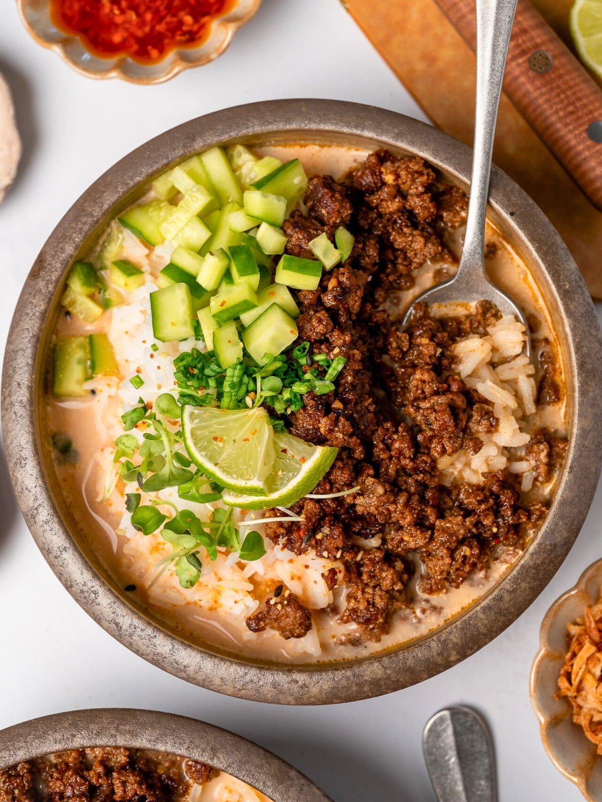 crispy beef and rice in a bowl with spicy miso ginger broth and sliced cucumber, chives, micro greens, and lime wedge garnishes
