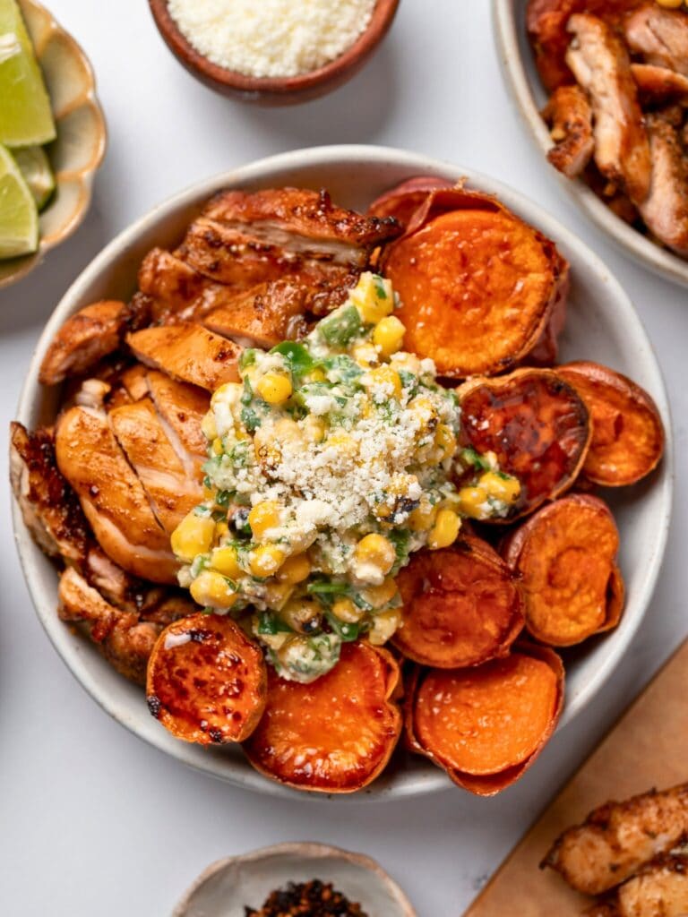 oven roasted sweet potato rounds and sliced Mexican chicken topped with street corn salad in a white bowl surrounded by garnishes