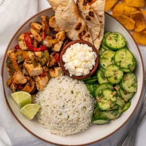a taco bowl with grilled chicken and peppers, cilantro lime rice, salsa macha cucumber salad, charred tortillas, queso fresco, and lime wedges