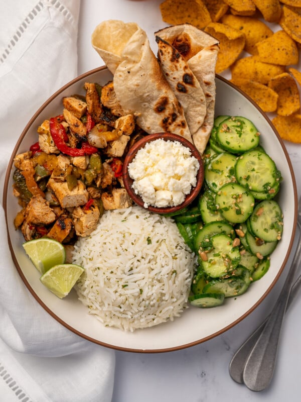 a taco bowl with grilled chicken and peppers, cilantro lime rice, salsa macha cucumber salad, charred tortillas, queso fresco, and lime wedges
