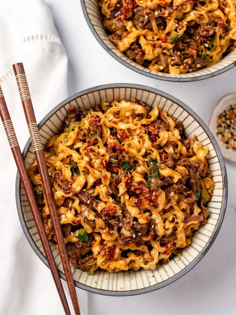 two bowls of beef noodles garnished with chili oil and toasted sesame seeds