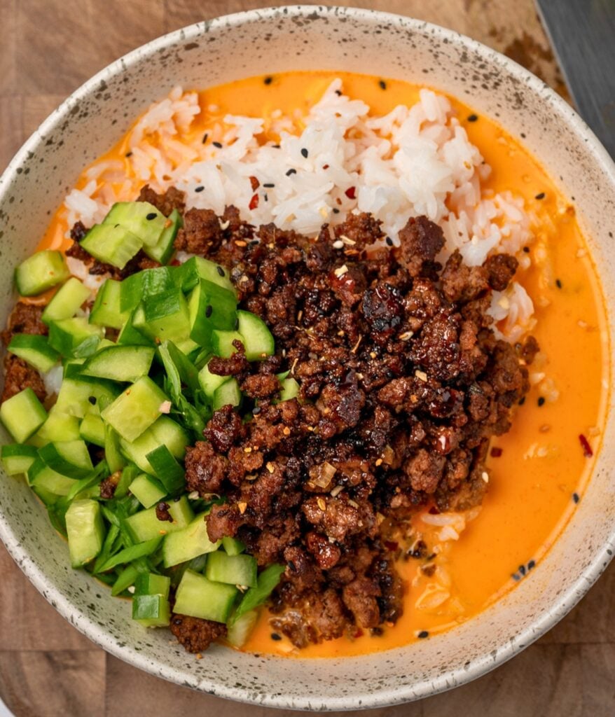 white bowl with gochujang broth, ground beef, rice, cucumbers, and green onions