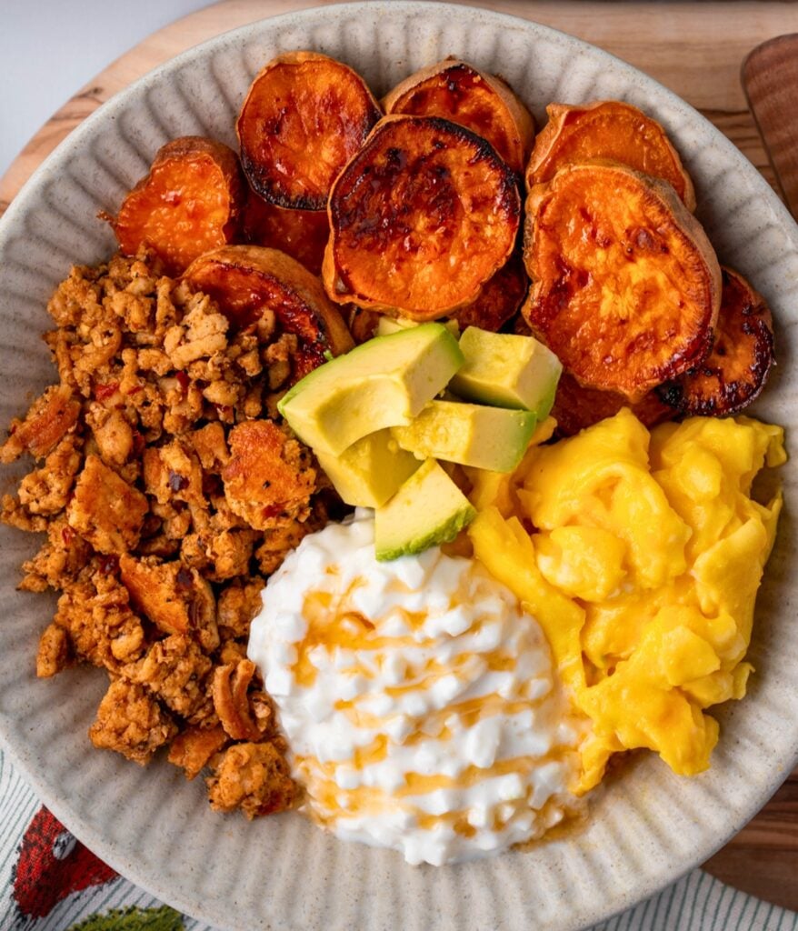 a bowl of roasted sweet potatoes, avocado, scrambled eggs, turkey breakfast sausage crumbles, and cottage cheese with honey