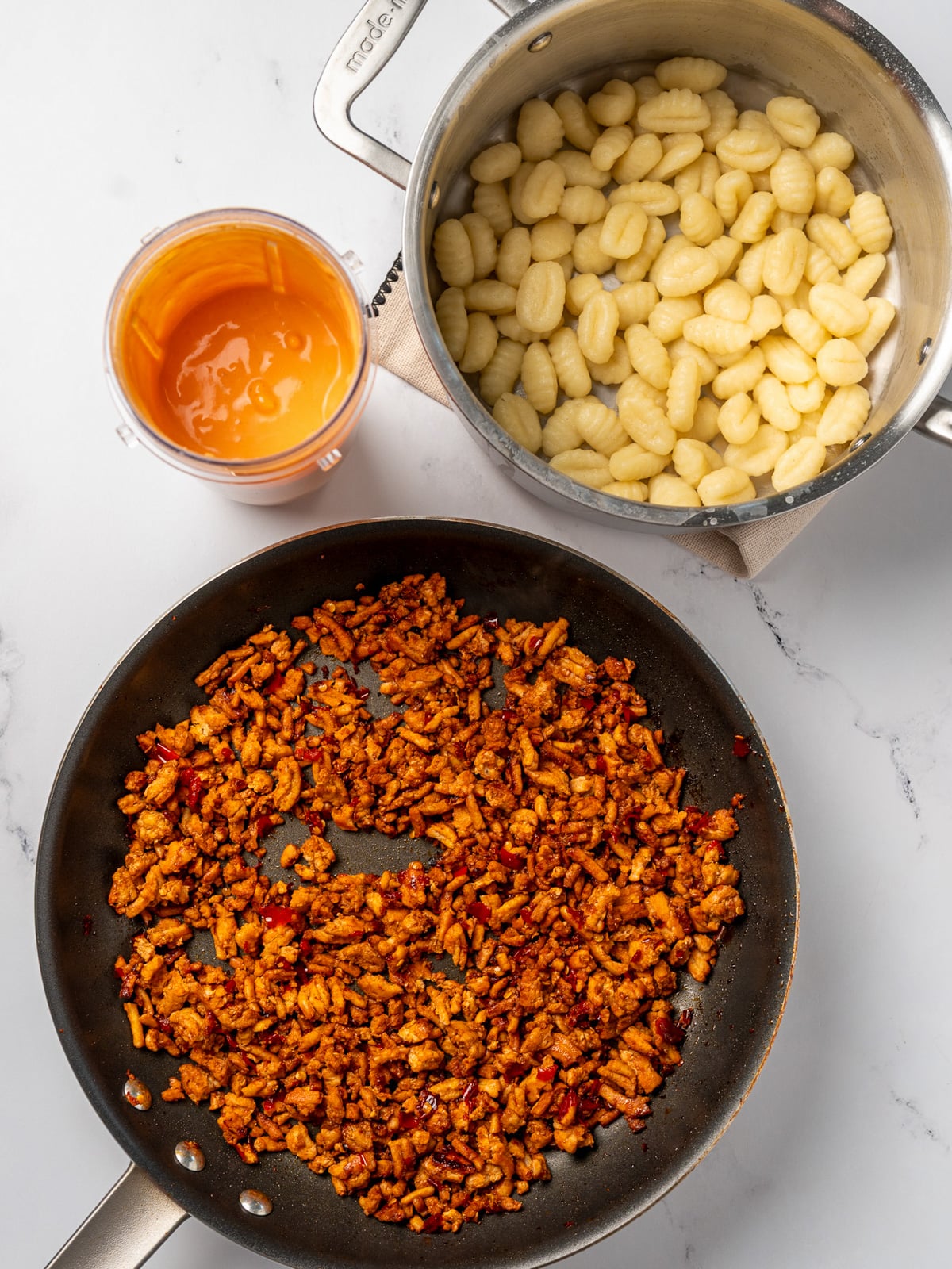 cooked chicken crumbles in a nonstick pan next to cooked potato gnocchi in a stainless steel pot and a blender cup with blended cottage cheese pasta sauce