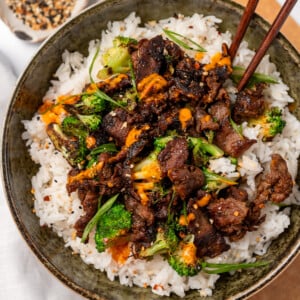 chopsticks in a bowl with steamed jasmine rice, black pepper beef, and broccoli garnished with sliced green onions, toasted sesame seeds, and sauce