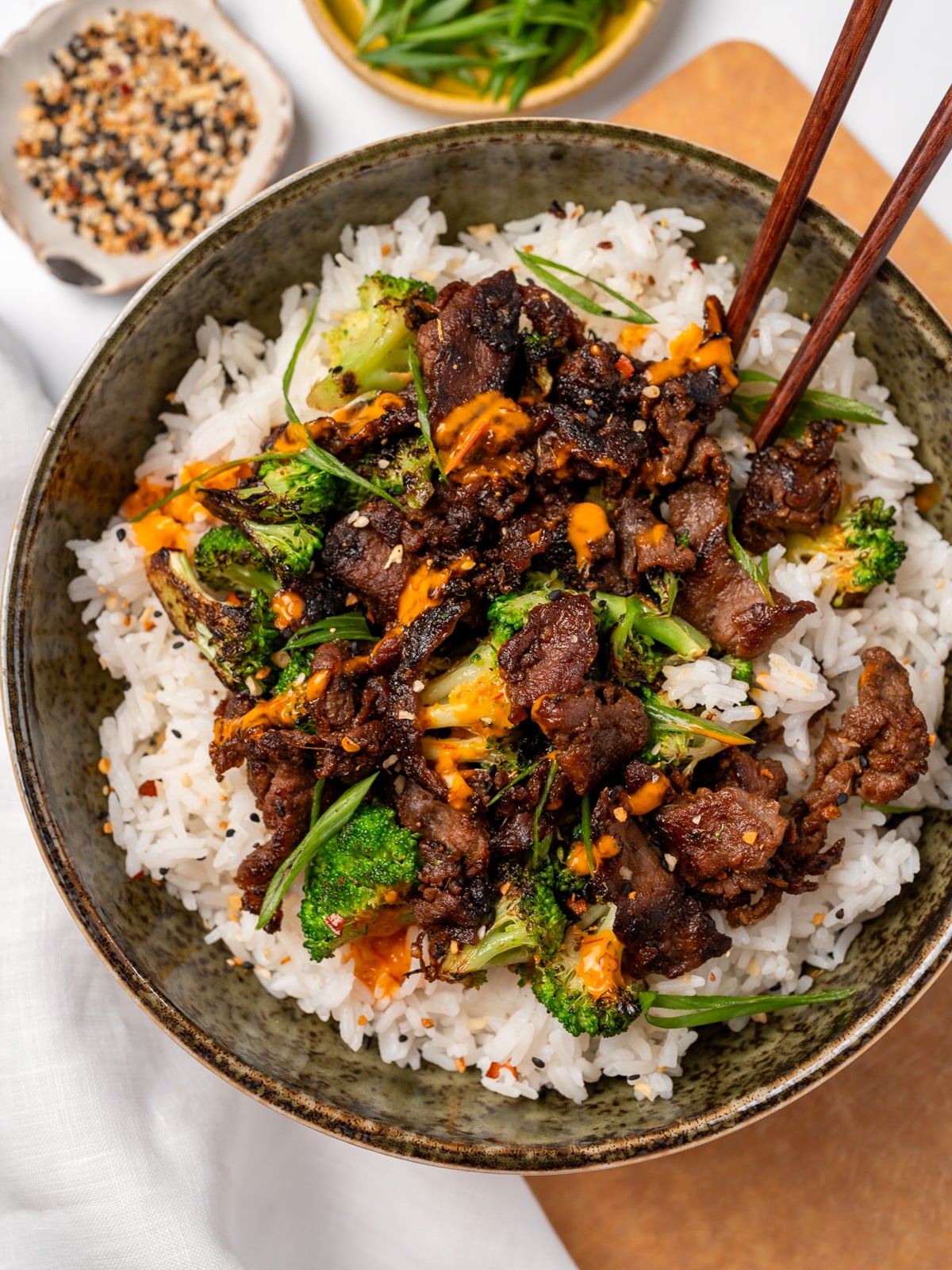 chopsticks in a bowl with steamed jasmine rice, black pepper beef, and broccoli garnished with sliced green onions, toasted sesame seeds, and sauce