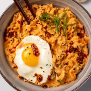 a bowl of creamy kimchi noodles garnished with chili oil, toasted sesame seeds, green onion, and a fried egg