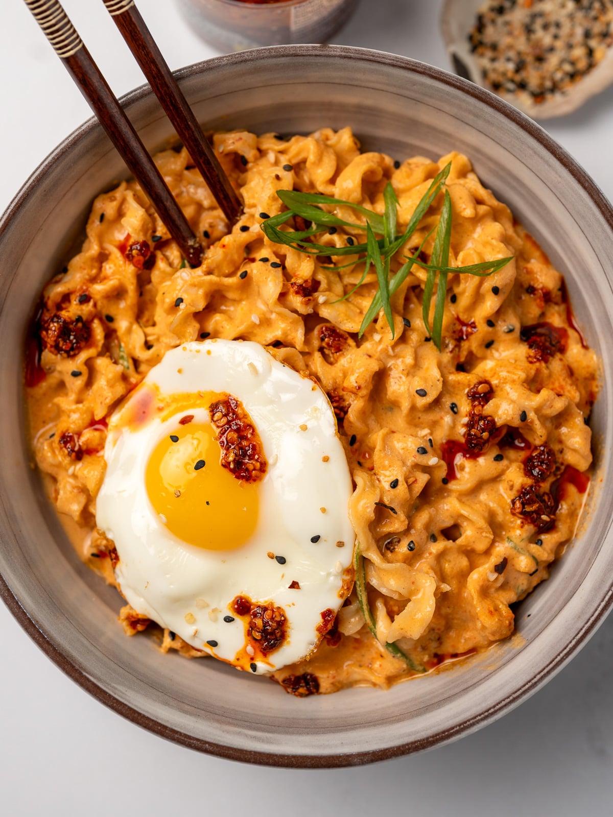 a bowl of creamy kimchi noodles garnished with chili oil, toasted sesame seeds, green onion, and a fried egg