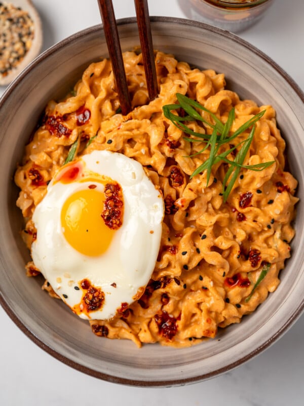 creamy kimchi noodles in a bowl with chopsticks and garnished with a fried egg, chili oil, toasted sesame seeds, and sliced green onion