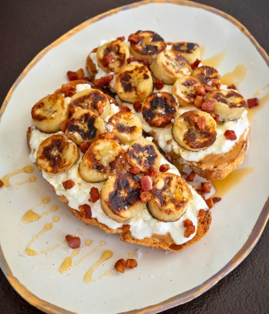 two slices of toast topped with caramelized banana slices, peanut butter, bacon, and honey