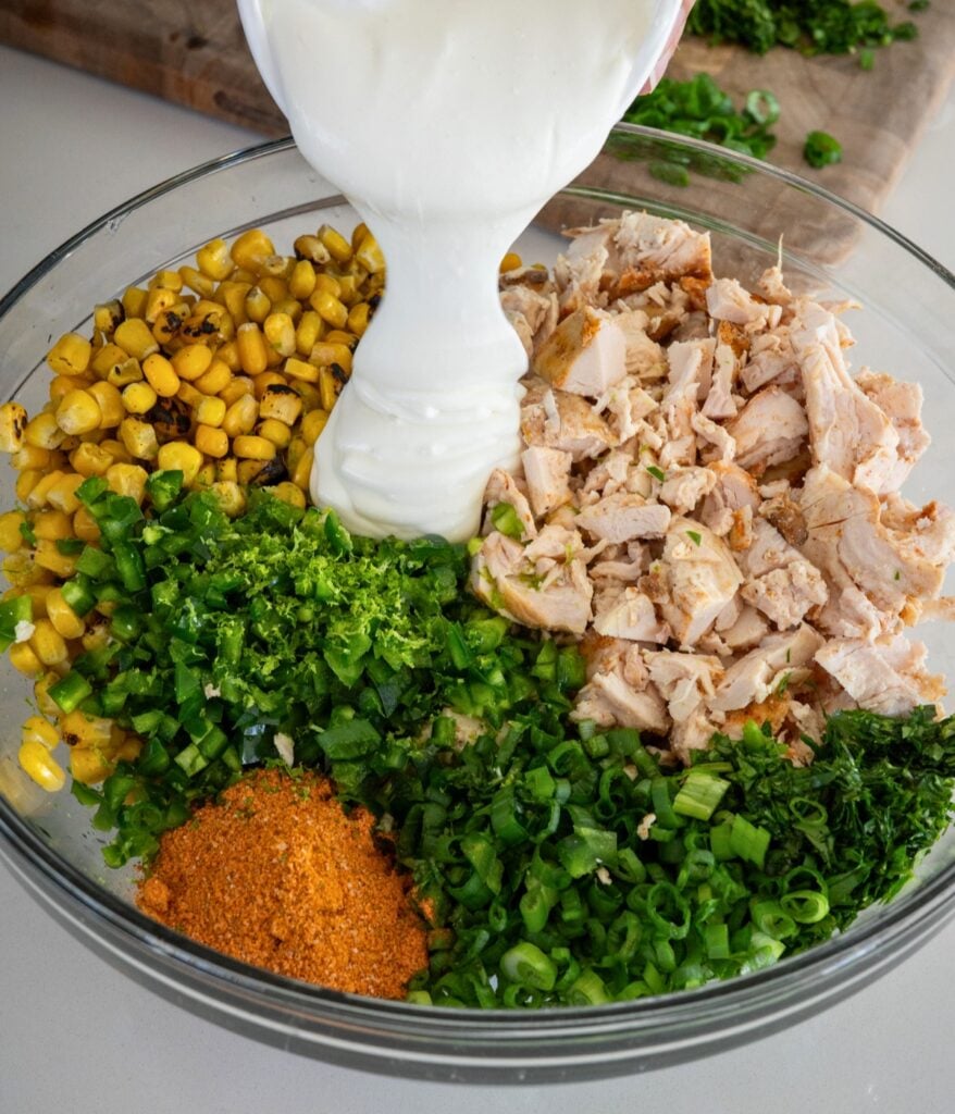 pouring blended cottage cheese into a bowl with elote seasoning, fire roasted corn, chopped chicken, and chopped herbs