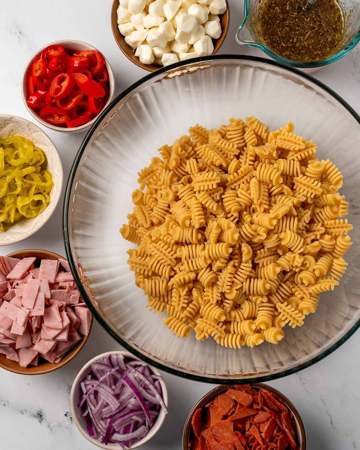 cooked pasta in a mixing bowl next to bowls of mozzarella pearls, dressing, pickled fresno peppers, pepperoncini, ham, red onion, and turkey peppperoni