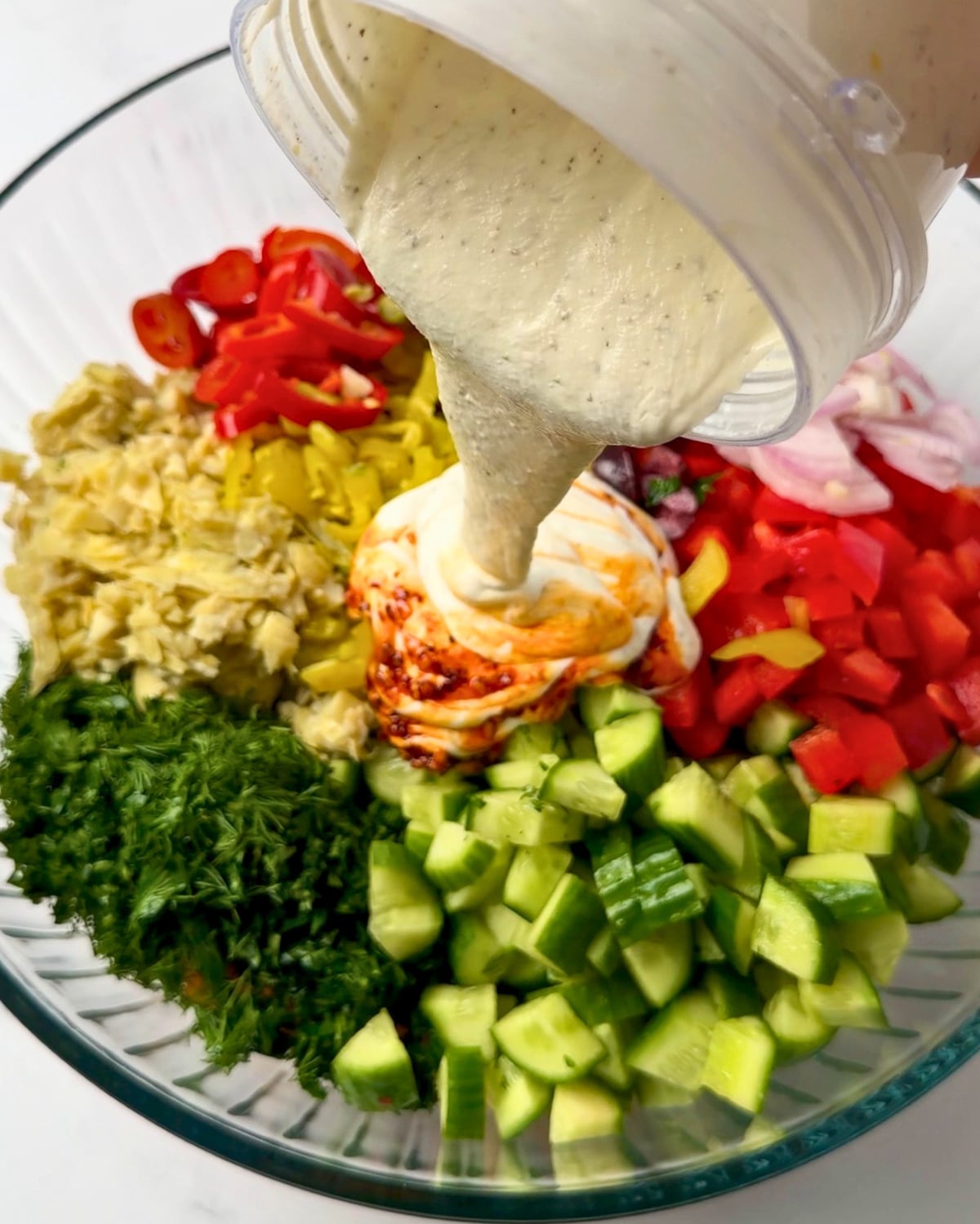 pouring feta sauce from a blender cup into a bowl of Greek pasta salad ingredients