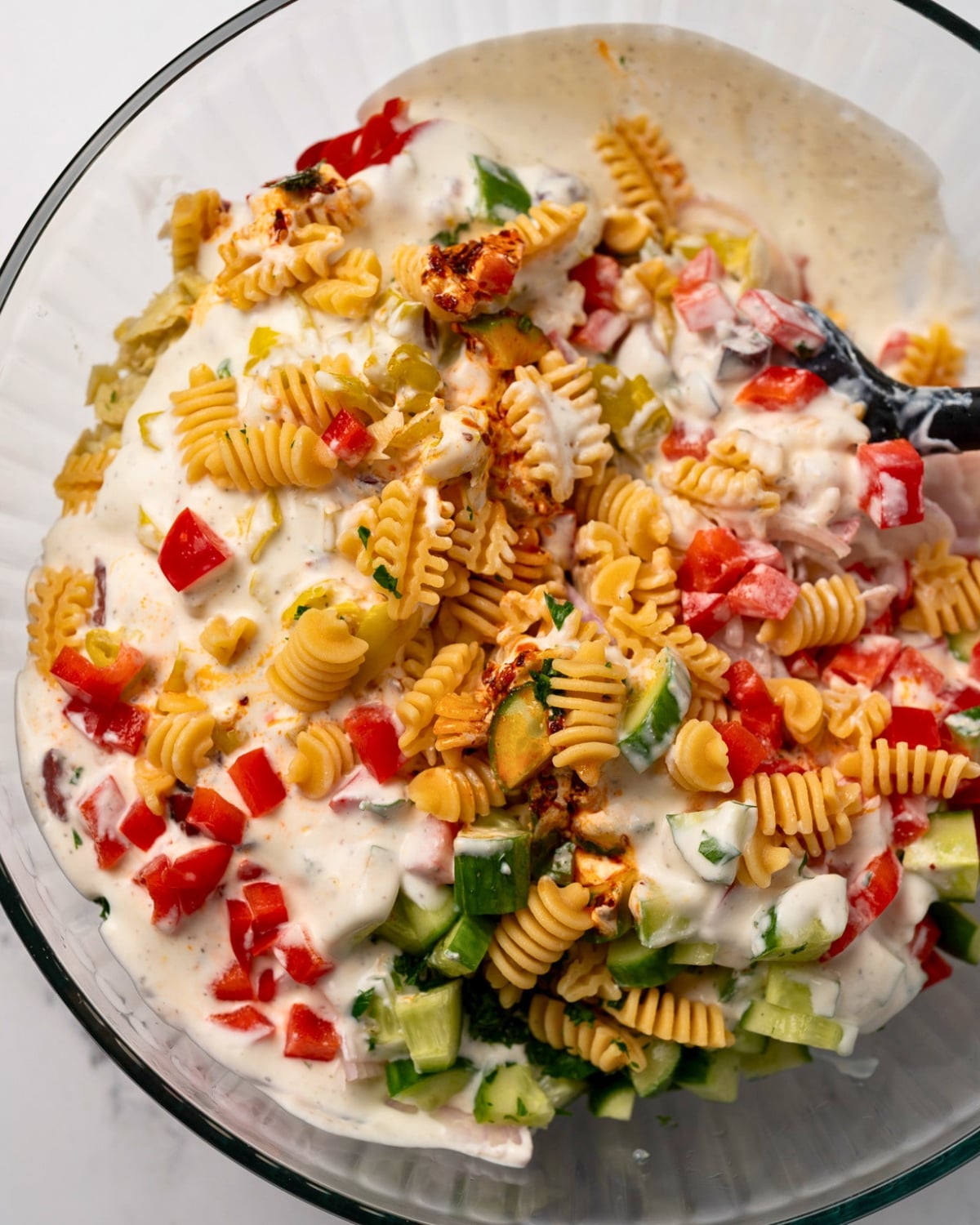 mixing cooked pasta and diced vegetables with a whipped feta sauce in a glass mixing bowl