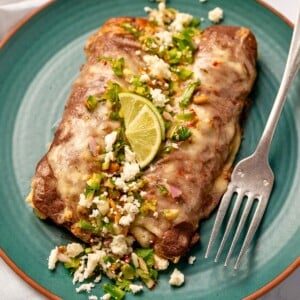two enfrijoladas on a green plate garnished with fresh jalape&ntilde;o salsa, queso fresco, and lime wedges