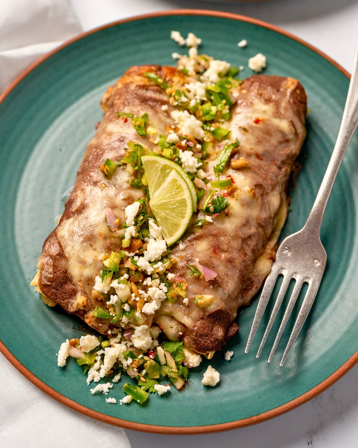 two enfrijoladas on a green plate garnished with fresh jalape&ntilde;o salsa, queso fresco, and lime wedges