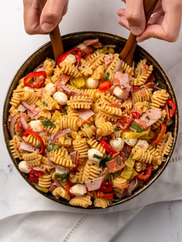 mixing deli style pasta salad together in a green bowl with two wooden spoons
