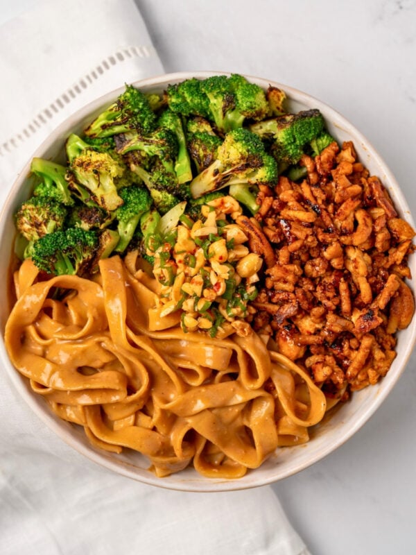 a white bowl with peanut butter noodles, chili honey ground chicken crumbles, pan roasted broccoli bites, and a chopped peanut scallion garnish