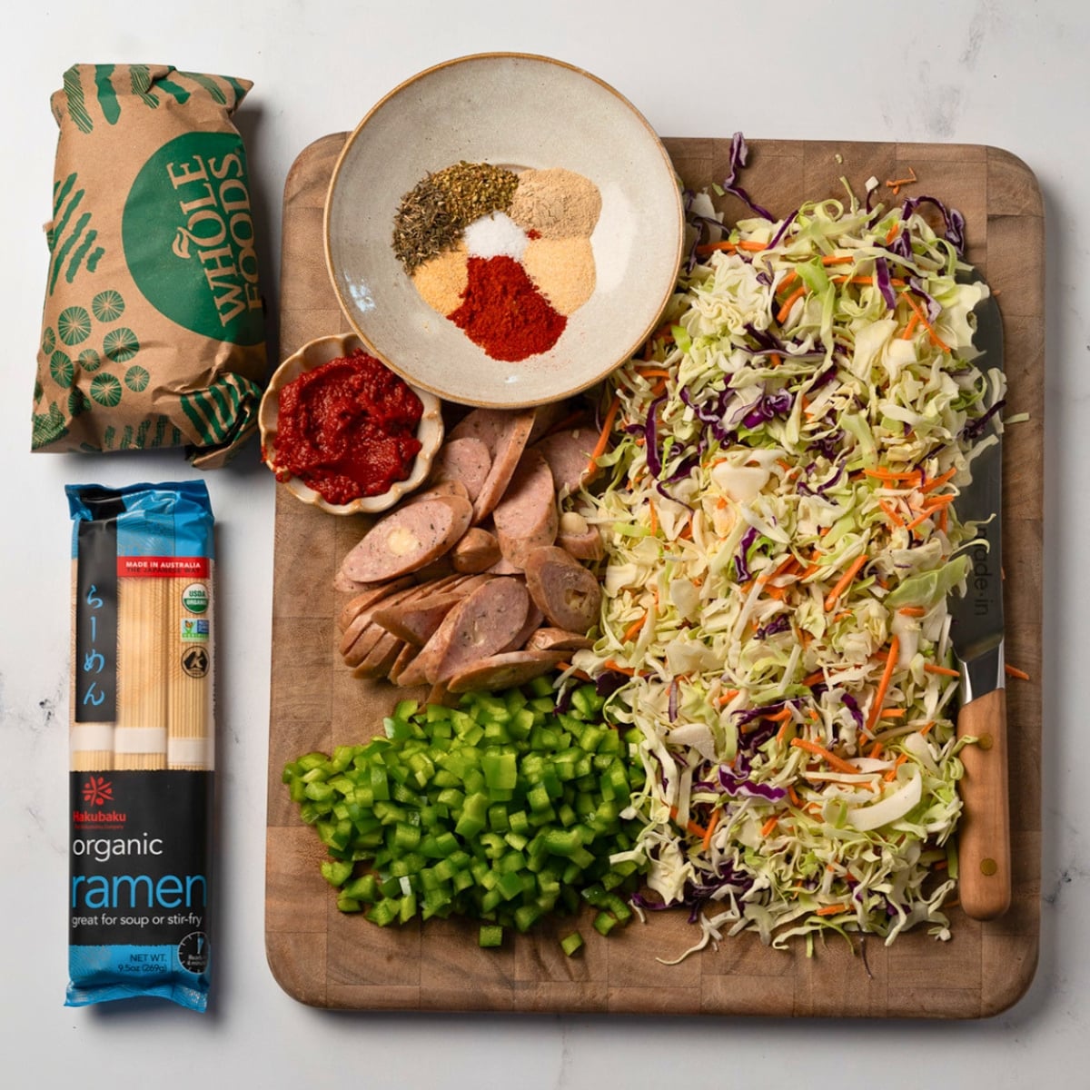 ground beef and ramen next to coleslaw mix, diced green bell pepper, sliced chicken sausages on a cutting board with a bowl of cajun seasoning blend and tomato paste
