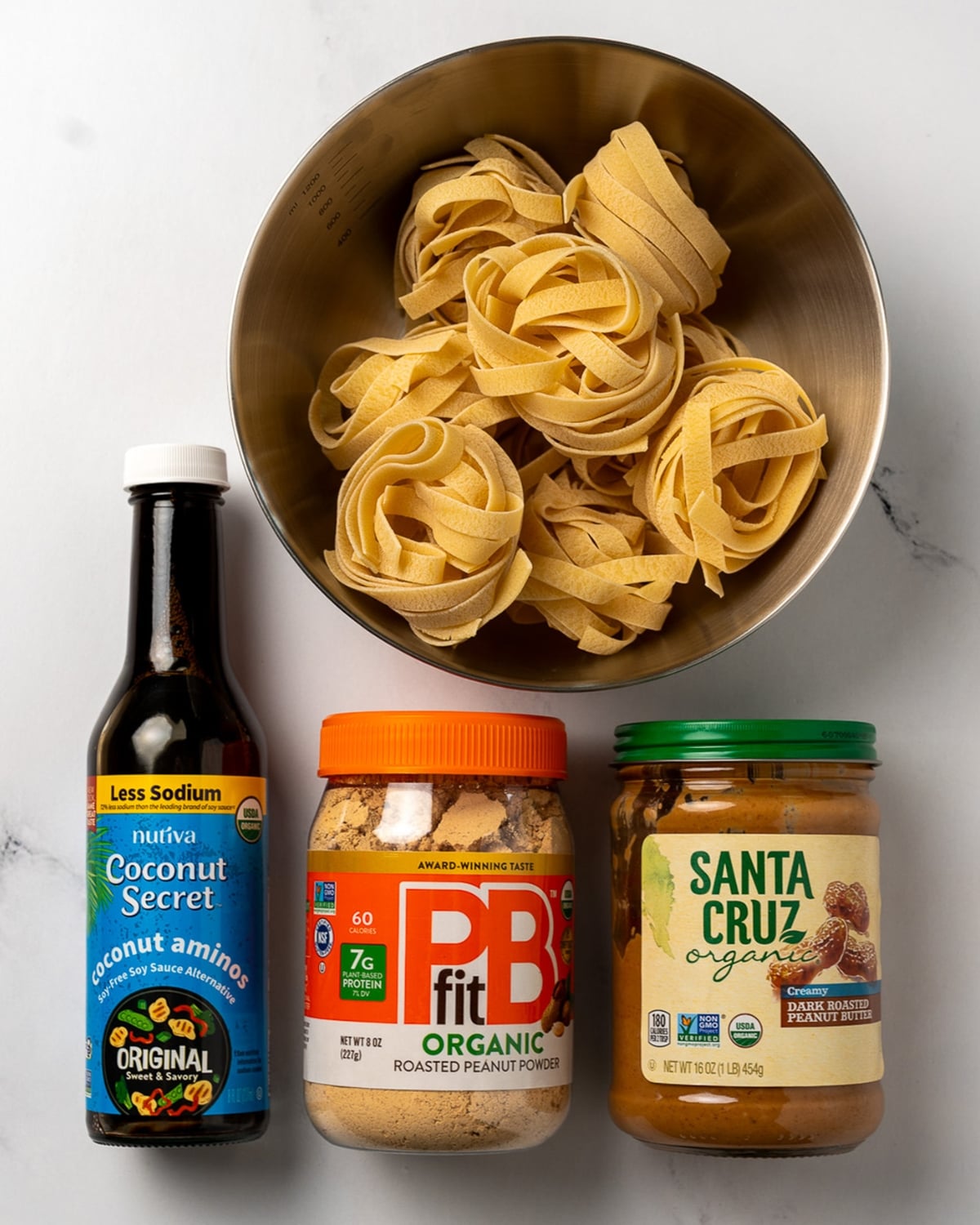 tagliatelle nests in a bowl next to a bottle of coconut aminos, powdered peanut butter, and peanut butter