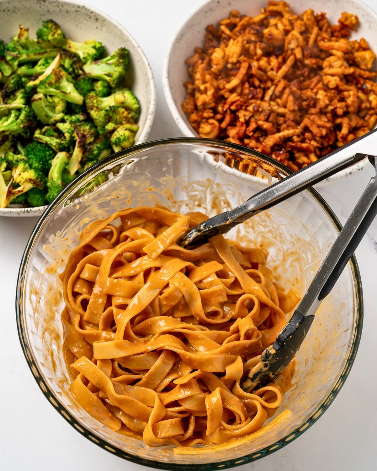 cooked pasta tossed in peanut sauce next to a bowl of chicken crumbles and a bowl of roasted broccoli