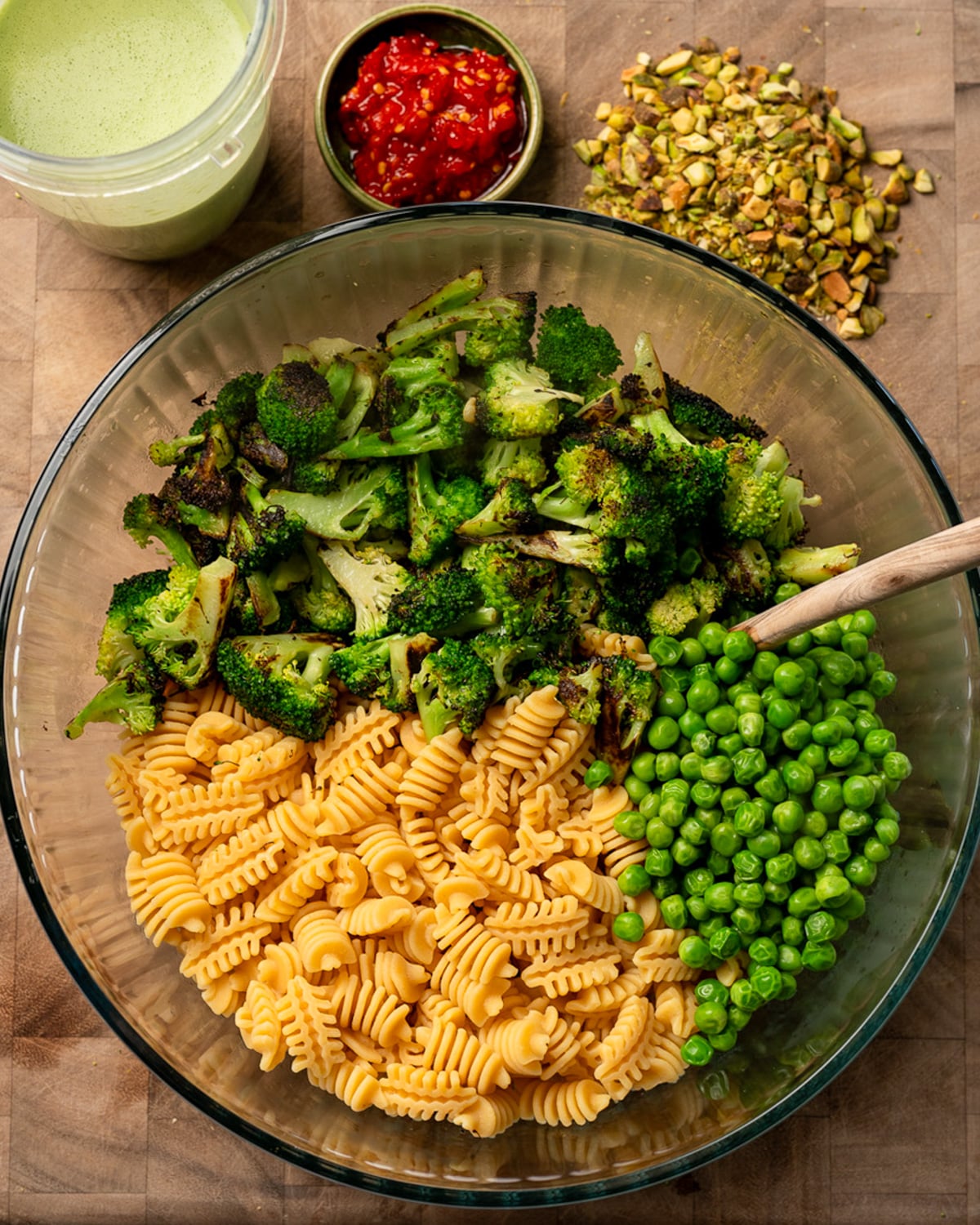 cooked pasta, roasted broccoli, and green peas in a mixing bowl next to a blender cup with cottage cheese pesto, a bowl of Calabrian chili peppers, and chopped pistachios