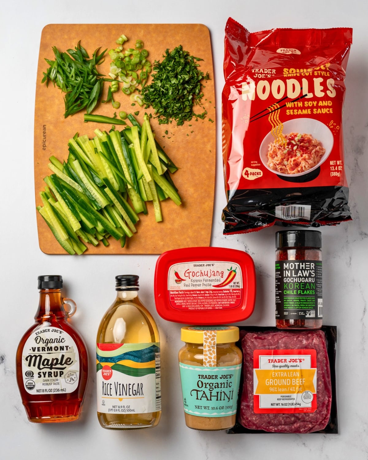 Trader Joe's instant noodles, maple syrup, rice vinegar, gochujang, tahini, and extra lean ground beef next to Korean chili flakes and a cutting board with cucumbers, green onion, and cilantro on it