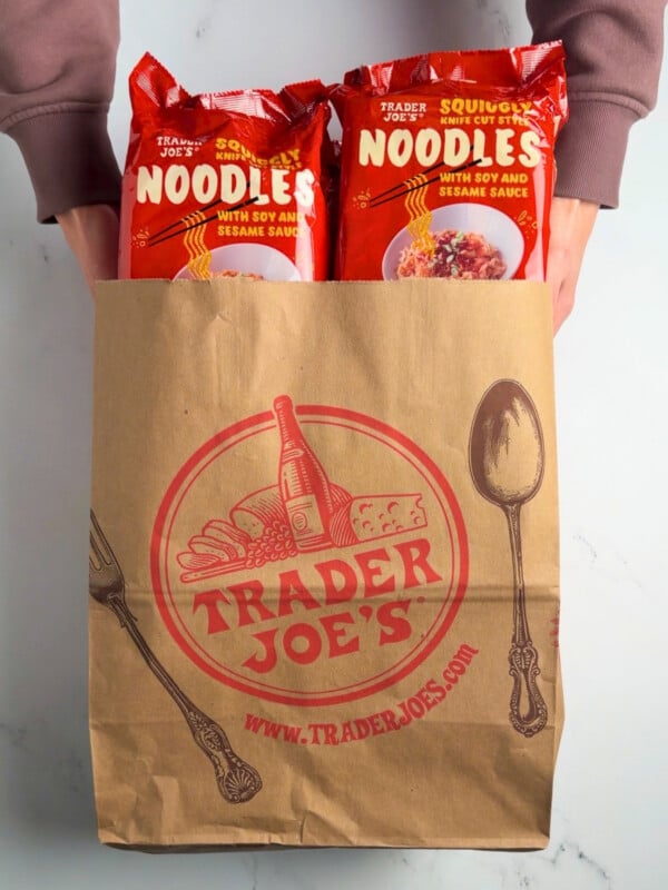 holding a paper Trader Joe's shopping bag filled with packages of squiggly knife cut style noodles