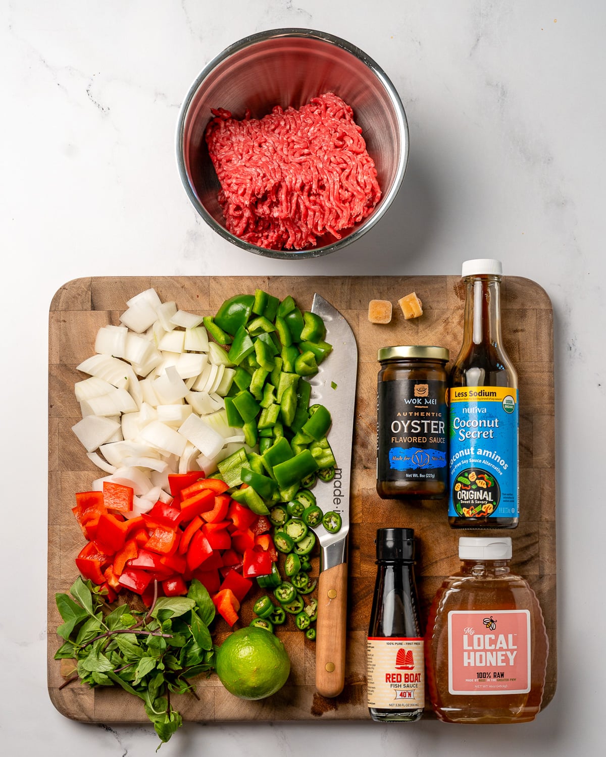 a bowl of lean ground beef next to a cutting board with diced onion and peppers, Thai basil, a lime, coconut aminos, honey, fish sauce, and oyster sauce