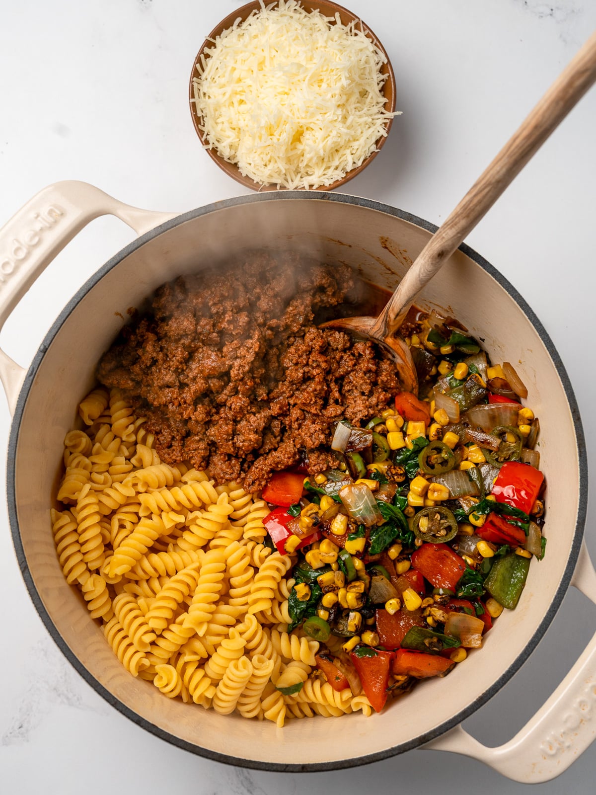 cooked pasta, taco beef, and saut&eacute;ed vegetables in a Dutch oven next to a bowl of asiago cheese