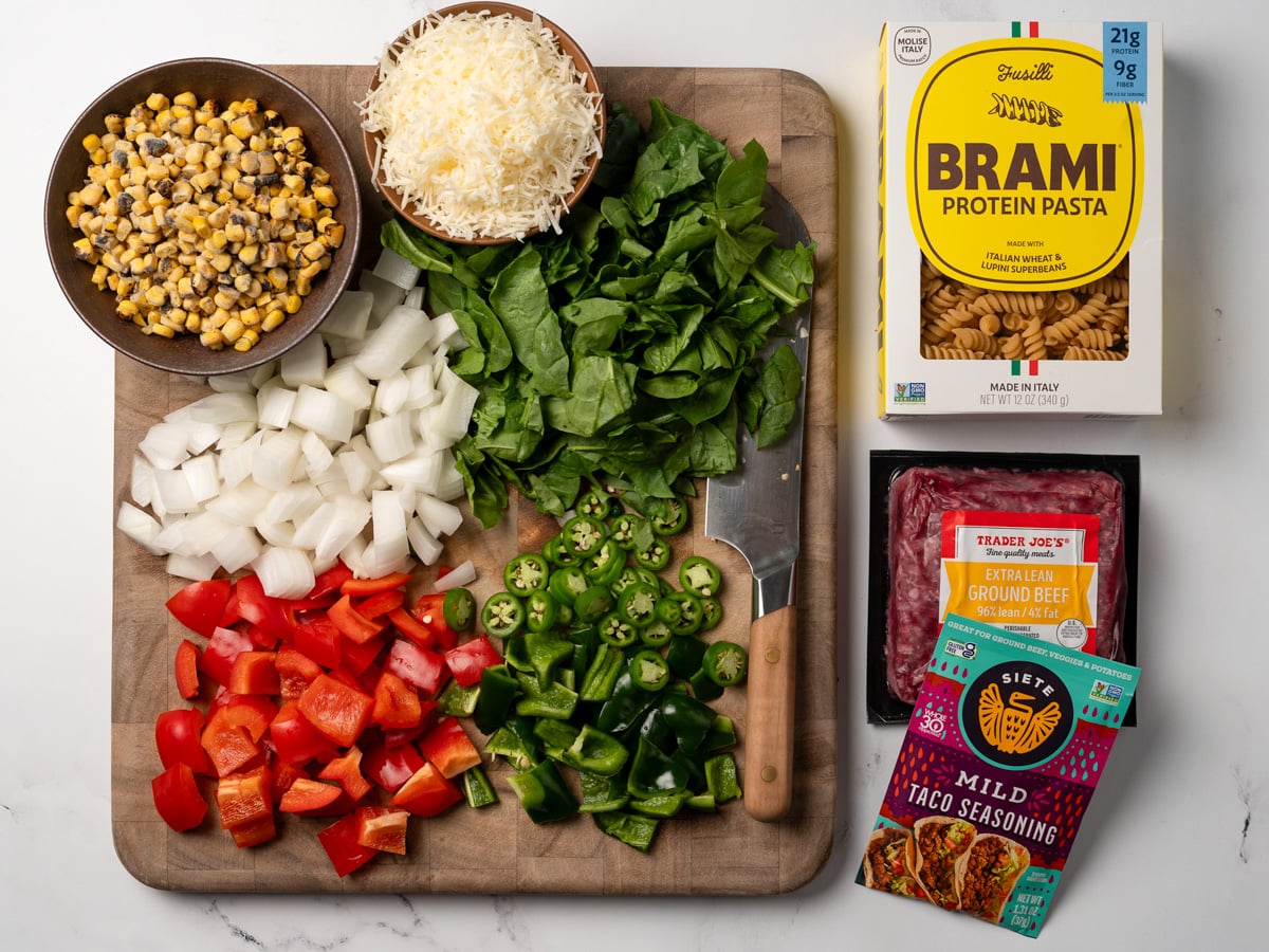 extra lean ground beef, taco seasoning, and brami protein pasta next to a cutting board holding diced peppers and onion, chopped baby spinach, and bowls of frozen fire roasted corn and grated asiago cheese