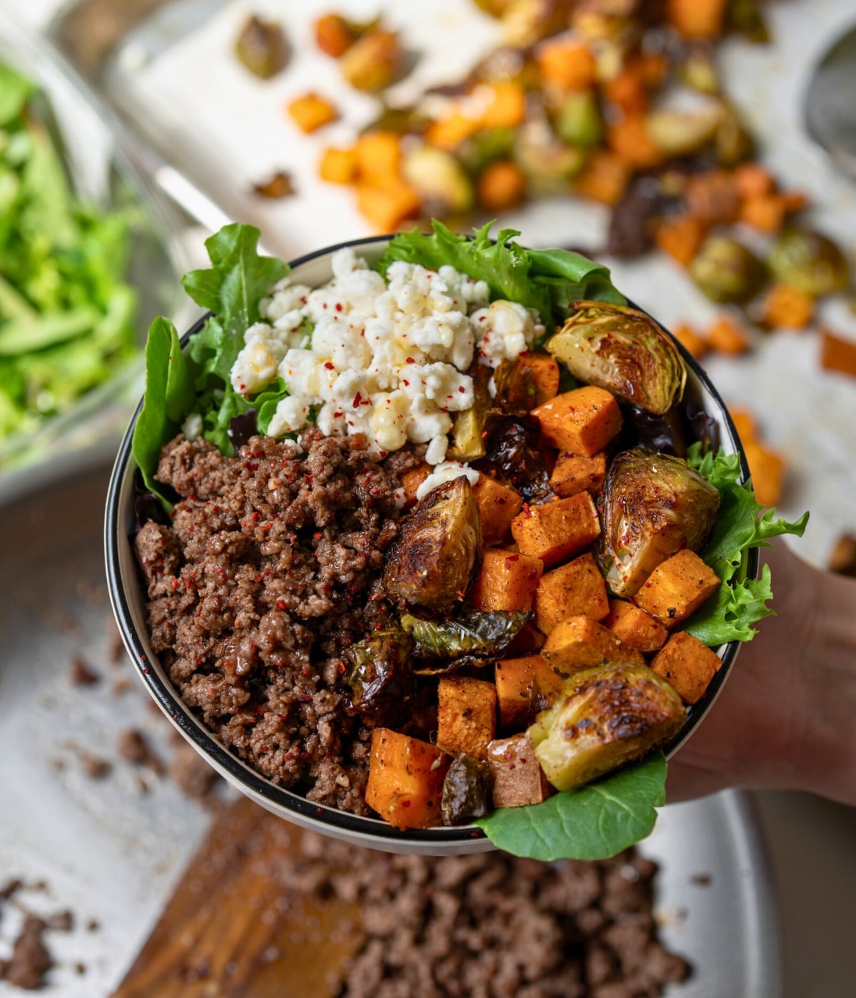 holding a bowl with ground beef, roasted vegetables, leafy greens, and cheese