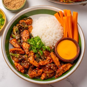 stir fried chicken breast and baby broccoli in a bowl with steamed rice, pickled carrots, sliced green onion, and sauce