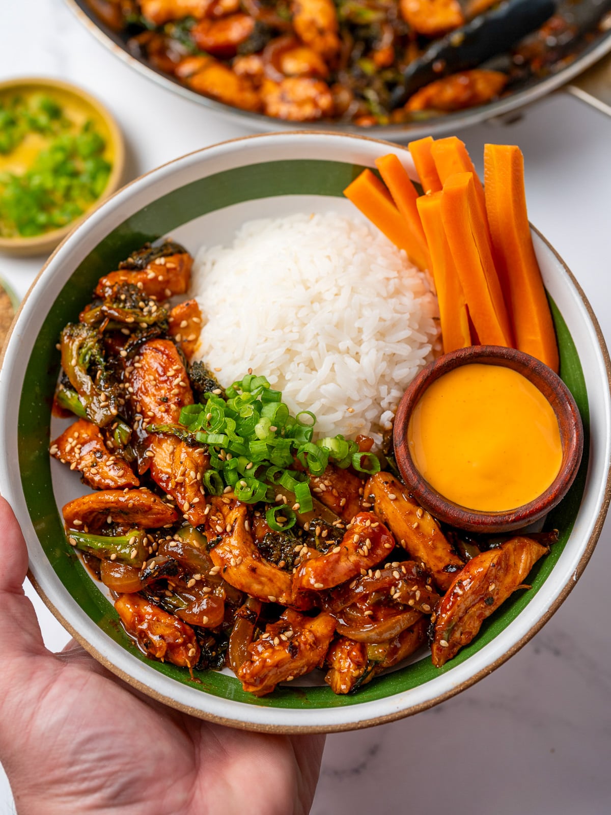holding a bowl with gochujang chicken breast and baby broccoli stir fry, jasmine rice, pickled carrots, sliced green onions, and a small bowl of sauce