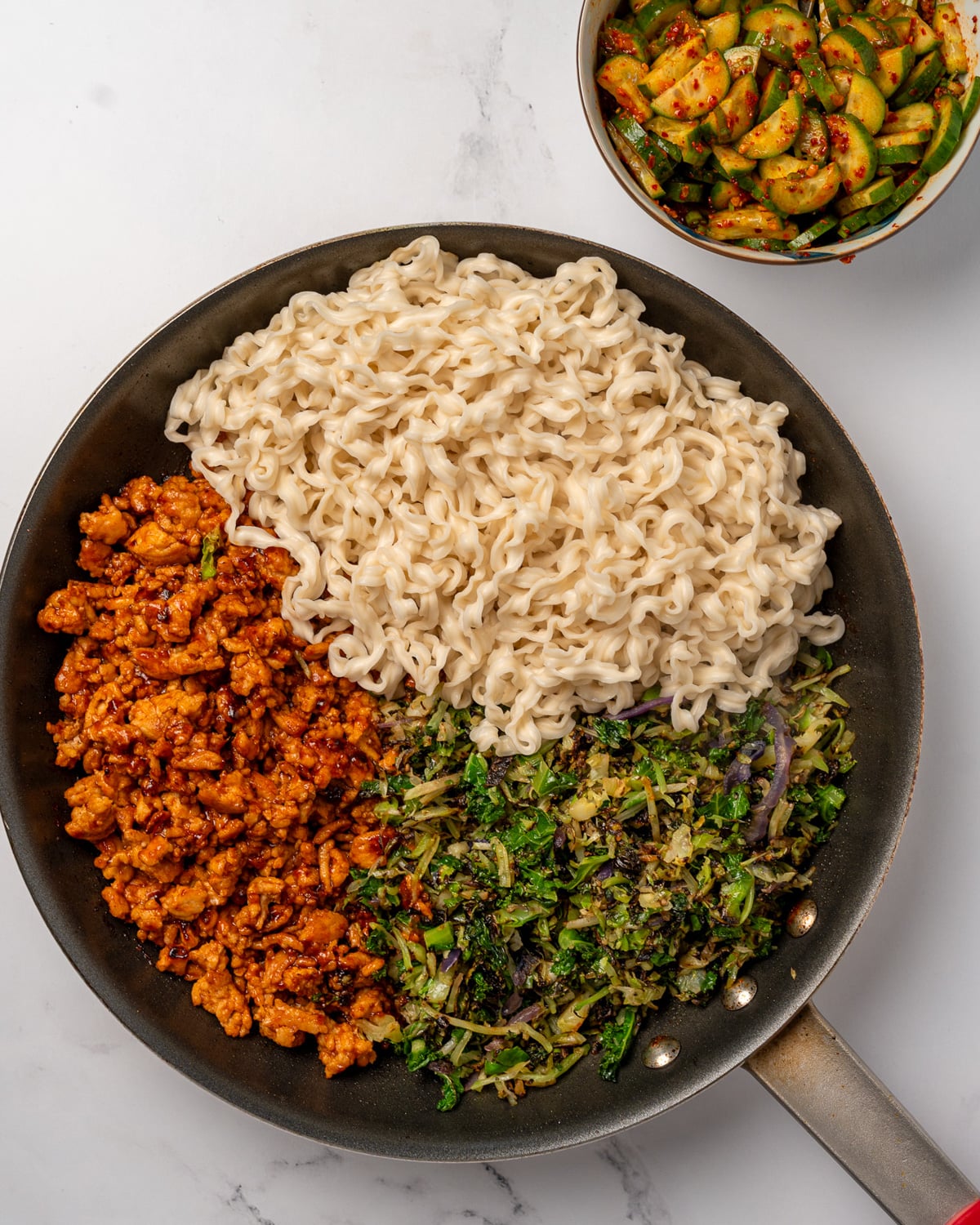 gochujang ground chicken, cruciferous crunch collection, and cooked noodles in a pan next to a bowl of pickled cucumbers