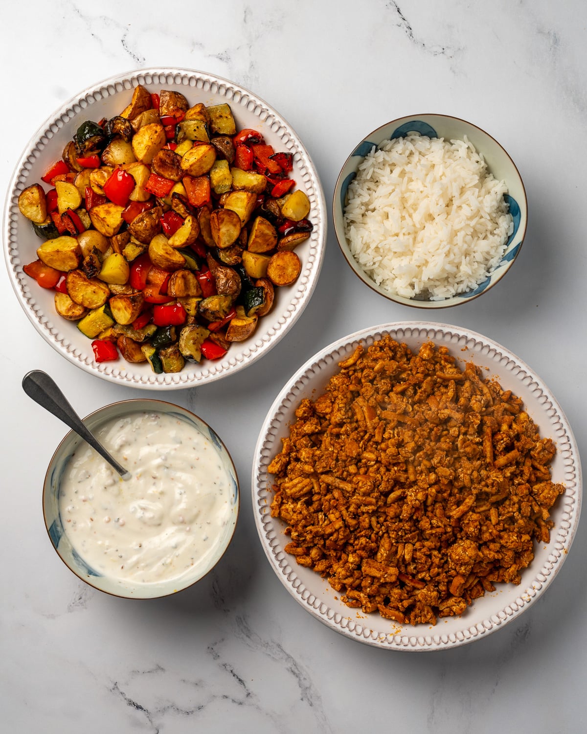 a bowl of roasted potatoes, zucchini, and peppers next to bowls of spiced chicken crumbles, steamed rice, and Greek yogurt