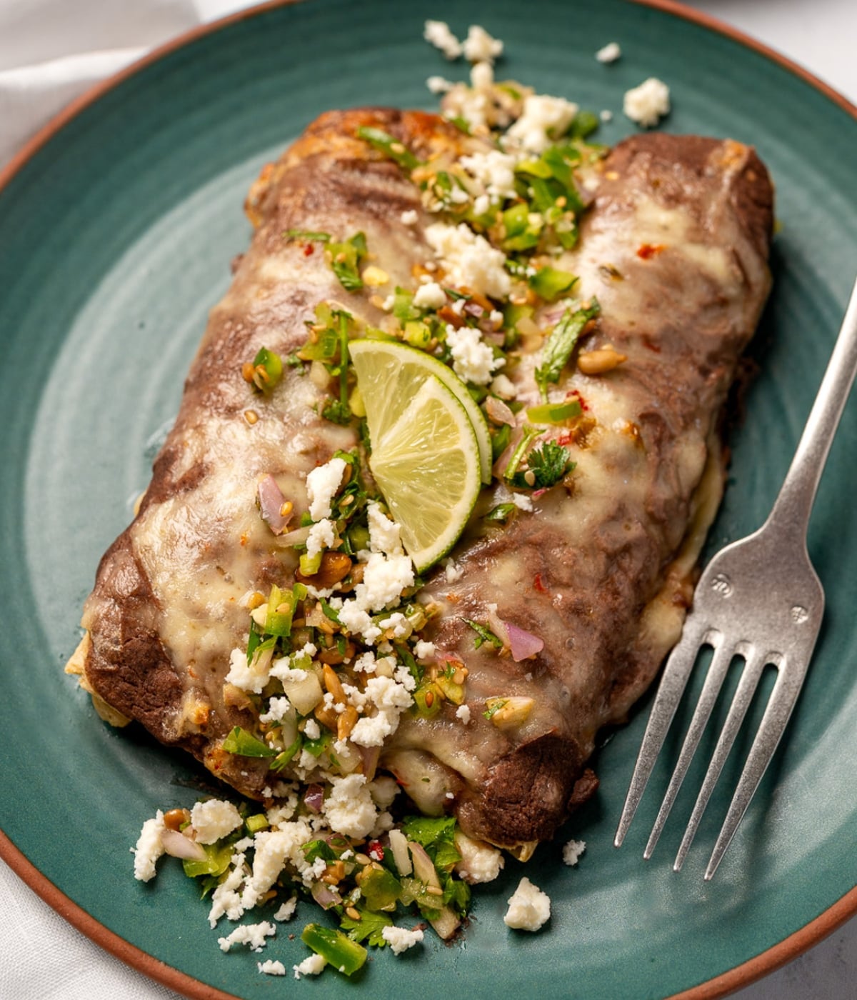 two enfrijoladas on a green plate garnished with lime wedges, cotija cheese, and jalapeño salsa